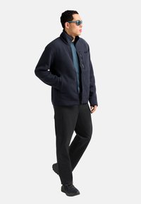 Giacca in pile a coste blu navy con colletto alto, tasca sul petto con zip e tasche laterali, abbinata a pantaloni cargo neri e scarpe da trekking.