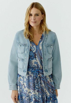 Femme aux cheveux blonds mi-longs portant une veste en jean bleu clair par-dessus une robe à imprimé floral paisley bleu et beige.