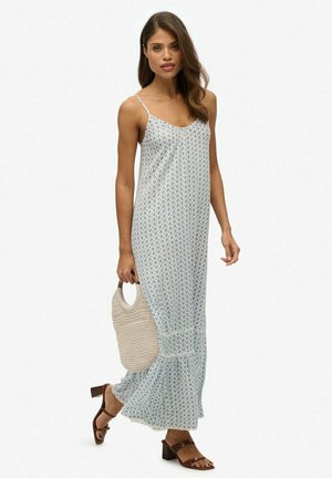 Helles blaues, blumenmuster Maxi-Kleid mit dünnen Trägern, weichem Stoff und gerüschtem Saum. Das Model hält eine gewebte beige Tasche und trägt braune Absatzsandalen.
