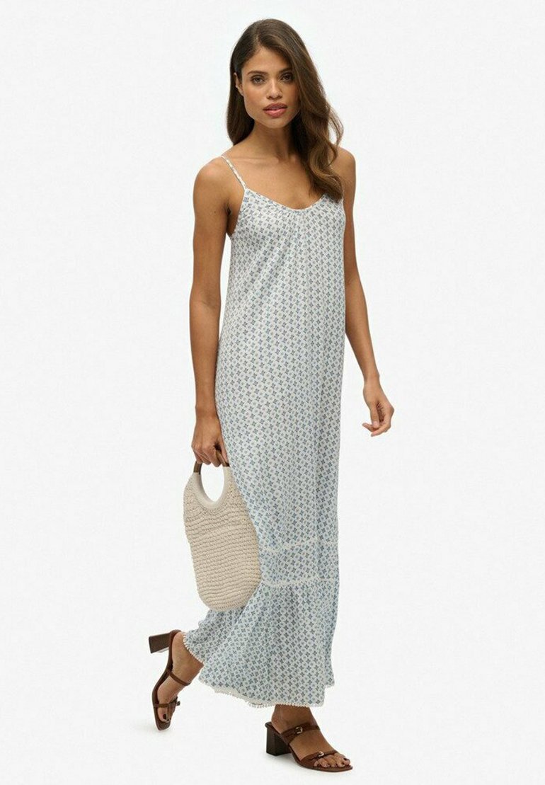 Helles blaues, blumenmuster Maxi-Kleid mit dünnen Trägern, weichem Stoff und gerüschtem Saum. Das Model hält eine gewebte beige Tasche und trägt braune Absatzsandalen.
