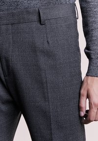 Primer plano de una persona que lleva puesto un pantalón de vestir texturizado gris oscuro y una camiseta de punto gris de manga larga, con la mano derecha relajada a un lado.