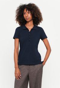 Marineblaues Polohemd mit kurzen Ärmeln, einem Kragen, einer Knopfleiste und glattem Stoff. Kombiniert mit hochgeschnittenen, strukturierten braunen Hosen.