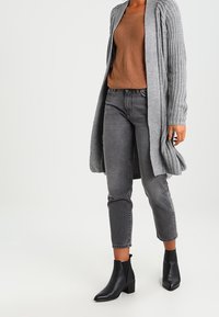 Šedý pletený kardigan přes hnědý žebrovaný svetr, doplněný šedými džíny s rovnými nohavicemi a černými kotníkovými botami s nízkým blokovým podpatkem.