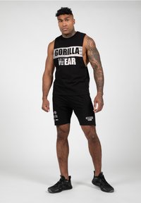 Zwarte mouwloze tanktop met "GORILLA WEAR" gedrukt op de borst, gecombineerd met zwarte shorts met logo's van het merk. Zwarte sportschoenen.