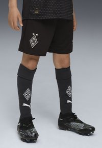 Kind, das schwarze Fußballshorts und Socken mit dem Logo von Borussia Mönchengladbach sowie schwarzen Puma-Sport schuhen auf grauem Hintergrund trägt.