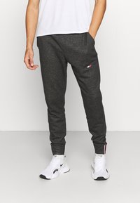 Graue Jogginghose mit schmal zulaufendem Design, featuring Seitentaschen und einem kleinen Logopatch am Oberschenkel. Kombiniert mit weißen Sneakers.