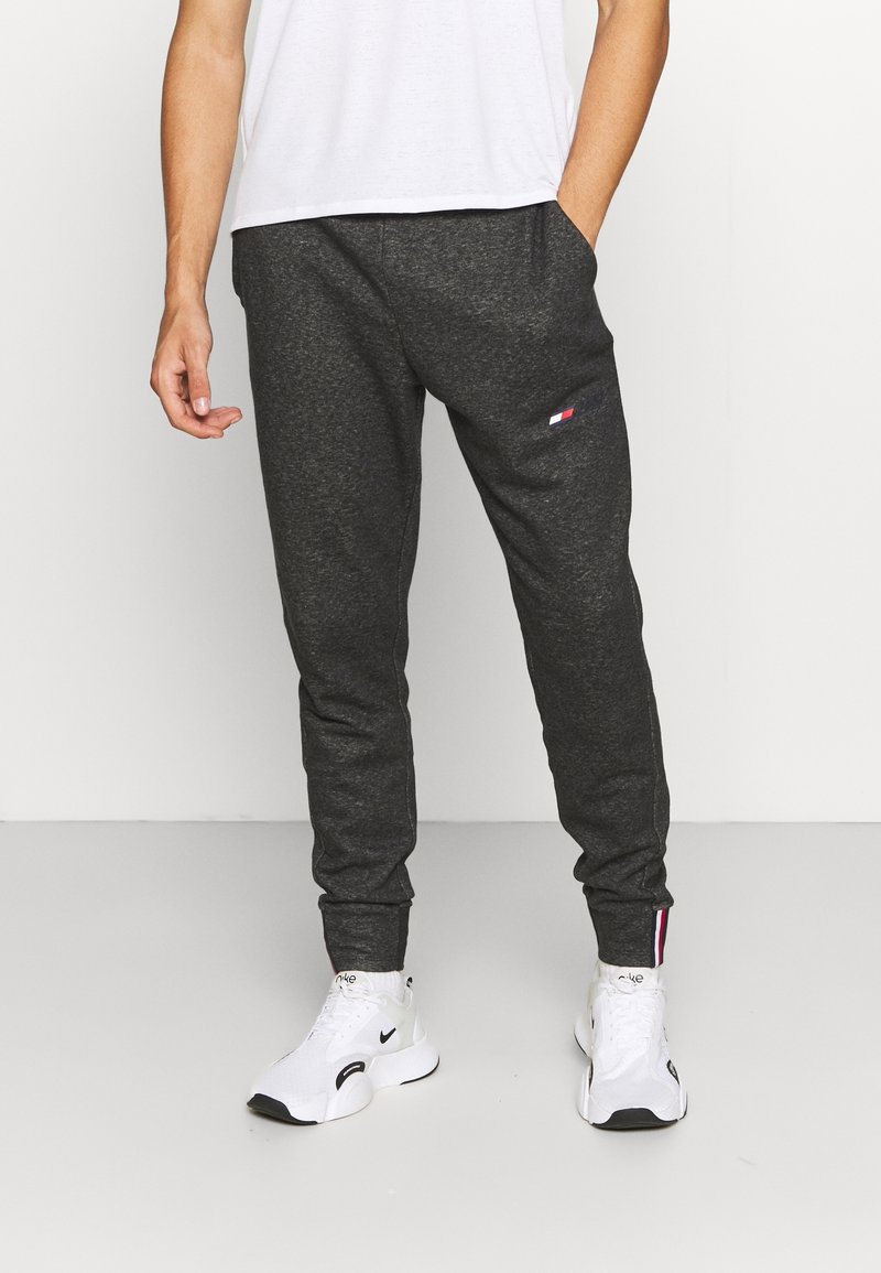 Graue Jogginghose mit schmal zulaufendem Design, featuring Seitentaschen und einem kleinen Logopatch am Oberschenkel. Kombiniert mit weißen Sneakers.
