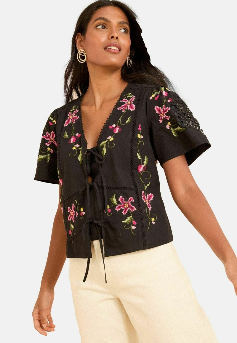 Melna blūze ar īsām piedurknēm, kas rotāta ar košiem ziedu izšuvumiem un V veida kakla izgriezumu. Priekšpusē ir saišķis, un uz piedurknēm ir smalka mežģīņu apdare.