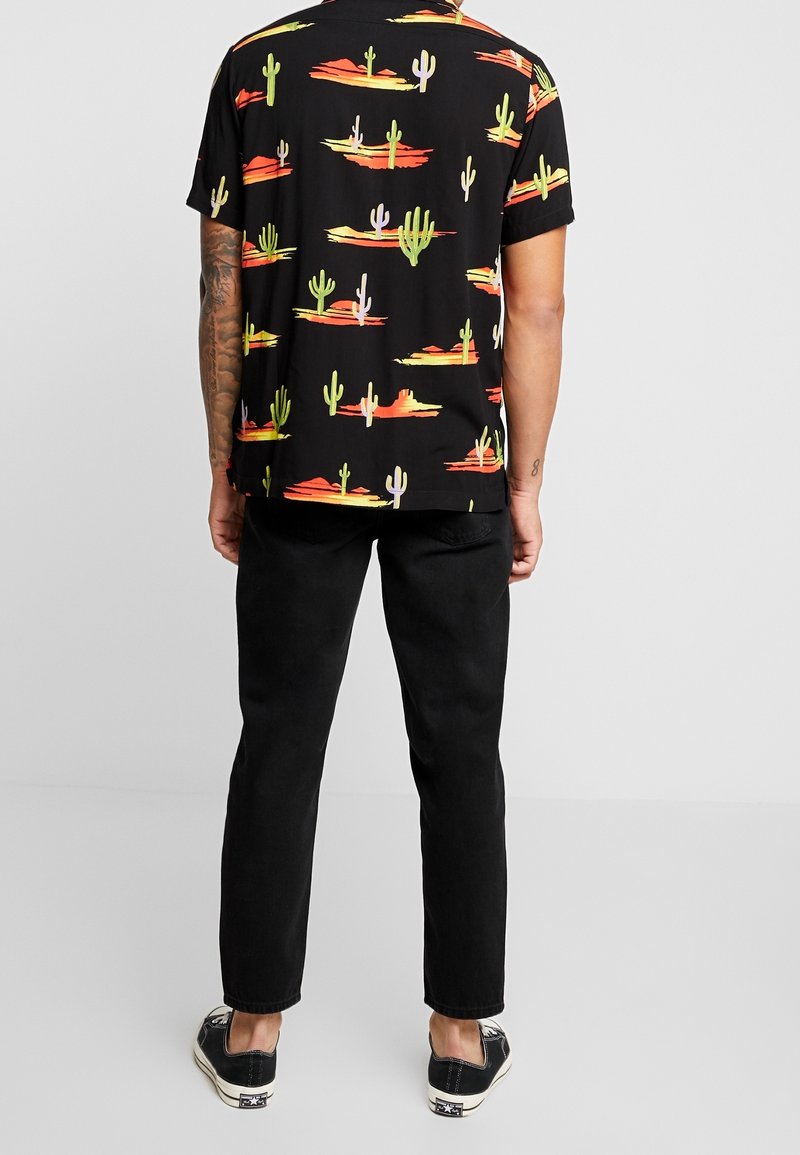 Homme portant une chemise noire à manches courtes avec un imprimé coloré de cactus et désert, un pantalon noir et des baskets basses noires, debout de dos.