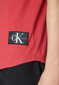 Camiseta de algodón roja con un parche rectangular negro que presenta el texto blanco "CK" y "Calvin Klein Jeans". Detalle de costura visible a lo largo del dobladillo.