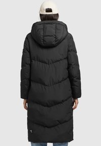 Donna che indossa un lungo cappotto imbottito nero con un grande cappuccio e un berretto in pile beige, posizionata su uno sfondo chiaro e semplice.