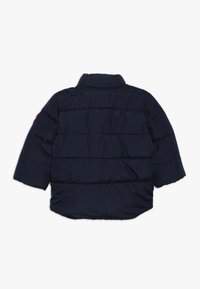 Marinblå puffjacka med hög krage, korta ärmar, vadderad yta och en rund röd patch på vänster axel.