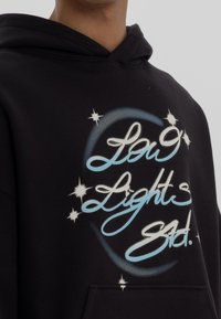 Sudadera de algodón negra con un diseño gráfico que incluye texto en cursiva en azul y blanco, rodeado de acentos estelares y un motivo circular.