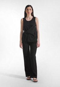 Musta hihaton toppi, jossa on solmittava yksityiskohta vyötäröllä, yhdistettynä väljien mustien housujen kanssa. Topissa on pyöreä kaula-aukko; molemmat vaatekappaleet ovat kangasta.