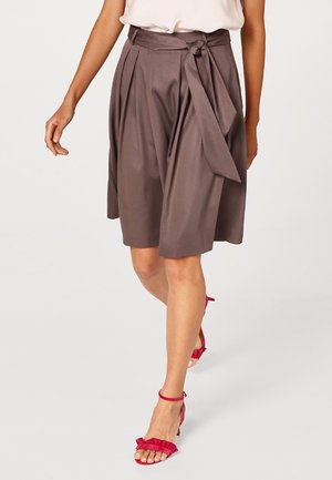 Woman wearing high-waist brown pleated skirt with tied belt and red open-toe heeled sandals walking forward on a plain background.