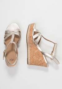 Metalické zlaté wedges sandály s korkovou základnou. Obsahují překřížené pásky a strukturovanou stélku. Zapínání na přezku na jedné straně.