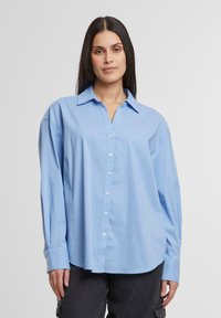 Camicia azzurro chiaro con colletto, maniche lunghe e vestibilità comoda. Il materiale sembra liscio e leggermente lucido. Sono visibili bottoni bianchi.