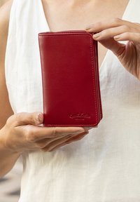 Červená kožená peňaženka s hladkou textúrou, prešitými okrajmi a vyleptaným logom. Držená v ruke, ukazujúca štíhly dizajn a vertikálnu orientáciu.