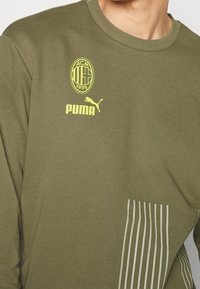 Chemise à manches longues vert olive avec logo circulaire jaune ACM et marque Puma. Présente des rayures verticales blanches sur le côté inférieur. Matière en coton.