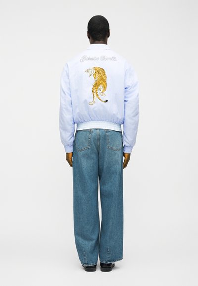 Homme de dos portant une veste bleu clair avec un tigre rugissant et le texte "Roberto Cavalli", un jean bleu ample et des chaussures noires sur fond blanc.