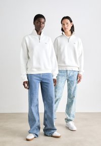 Sudadera blanca con cremallera y puños acanalados, combinada con jeans de pierna ancha azul claro. Uno de los jeans presenta un patrón sutil, con zapatillas blancas.