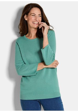 Glunderende vrouw met bruin haar, gekleed in een gestructureerde groene langwerpige trui en blauwe spijkerbroek, staande tegen een lichte achtergrond.