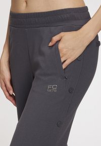 Gray athletic pants featuring an elastic waistband, side pockets, and decorative buttons along the leg. Embroidered logo on the thigh.