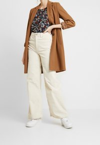 Brązowy długi płaszcz na kwiecistej granatowej bluzce i kremowych szerokich spodniach z kordurojowym materiałem, w połączeniu z białymi sneakersami.