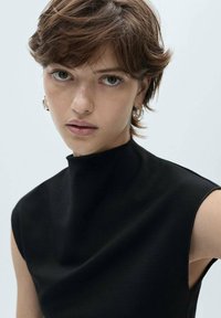 Top senza maniche nero con collo alto, realizzato in tessuto morbido. Il modello presenta pelle chiara e capelli corti castani con orecchini a cerchio.
