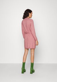 Robe tricotée rose pâle avec des manches longues, taille cintrée et ourlet côtelé. Le mannequin porte des bottines en daim vert avec des talons carrés.