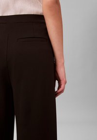 Nahaufnahme der Taille und Hand einer Person, die schwarze, maßgeschneiderte Hosen mit einer Gesäßpaspeltasche und einem hellbeigen Oberteil trägt.