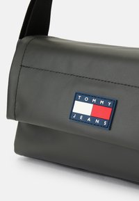 Pilkas poliesterio maišas su lygiu paviršiumi, turintis sukurtą viršų ir mėlyną bei raudoną prekės ženklo etiketes "TOMMY JEANS". Juodas petnešų diržas.
