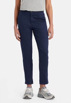 Osoba nosząca granatowe spodnie typu slim fit, szare casualowe sneakersy oraz jasnoszary top, stojąca na białym tle.