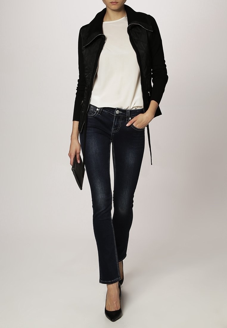 Femme portant une veste noire, une chemise blanche, un jean bleu foncé, des talons hauts noirs, tenant une pochette noire en marchant.