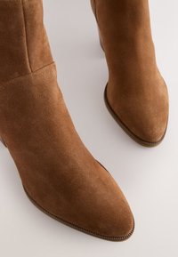 Paar braune Wildleder-Stiefeletten mit spitz zulaufender Zehenpartie und flachen Absätzen auf hellem Hintergrund.
