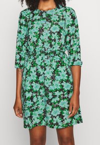 Robe à fleurs avec un fond noir, présentant des fleurs bleu clair et des feuilles vertes ; taille cintrée et manches trois-quarts.