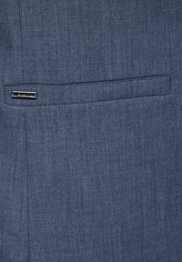 Tessuto di un blazer blu scuro con una sottile trama. Presenta una piccola tasca orizzontale e un elegante etichetta metallica vicino alla tasca.