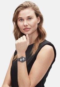 Schwarze Uhr mit einem runden grauen Zifferblatt, strukturiertem Armband, minimalen Markierungen und einer kleinen Krone. Der Träger hat einen neutralen Gesichtsausdruck.