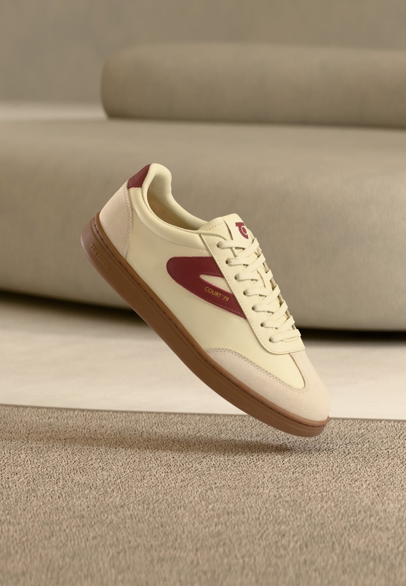 Krémovo vínová kožená teniska s gumovou podrážkou, označená "COURT 79", vznáša sa nad textúrovaným zelenohnedým kobercom s pozadím béžovej pohovky.