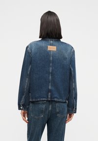 Personne aux cheveux noirs mi-longs portant une veste en jean bleue et un jean, vue de dos sur un fond blanc uni.