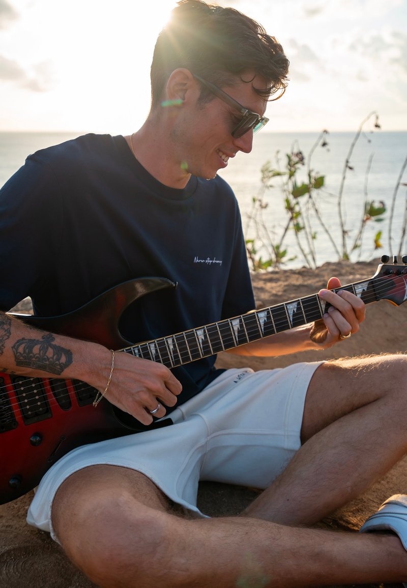 Čierna a červená elektrická gitara, ktorú drží osoba v tmavom tričku a bielych šortkách, sediaca na piesku s oceánom a slnkom v pozadí.