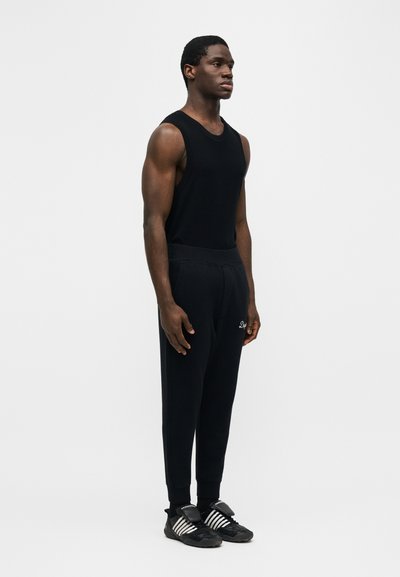 Άνδρας που στέκεται πλαγίως, φοράει μαύρο αμάνικο τοπ, μαύρο παντελόνι με λευκό λογότυπο και μαύρα αθλητικά παπούτσια με λευκές ρίγες.