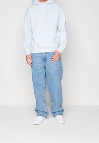 Ljusblå huvtröja med en framficka och vitt logo, i kombination med löst sittande ljusblå jeans och vita sneakers.