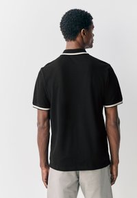 Musta poolopaita, jossa valkoinen reunus, lyhyet hihat ja klassinen kaulus, valmistettu sileästä kankaasta, istuva malli, ilman näkyviä logoja.