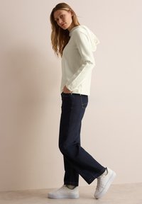 Sudadera de manga larga en color marfil con capucha, que presenta un detalle de "Gracias", combinada con pantalones de mezclilla oscuros y zapatillas blancas.