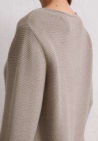 Beige gebreide trui met een gestructureerd, ribbelpatroon, ronde halslijn en een relaxte pasvorm, zijwaarts weergegeven om het ontwerp te benadrukken.