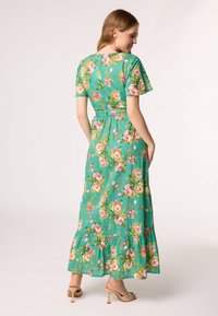 Flohendes Maxikleid in Grün, mit kurzen Ärmeln, tailliertem Schnitt und Rüschensaum. Das Blumenmuster enthält Rosen in Rosa- und Gelbtönen.