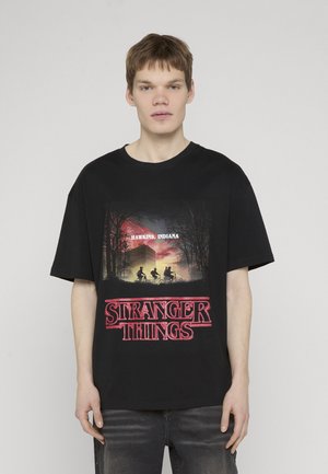 Joven con una camiseta negra de "Stranger Things" con siluetas en bicicletas, árboles oscuros y texto "Hawkins, Indiana" contra un cielo al atardecer.