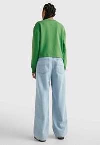 Osoba s copánky, oblečená v zeleném triku s dlouhým rukávem a světlých džínách s širokými nohavicemi, stojí před jednoduchým pozadím.