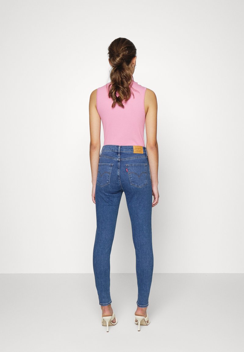 Hautenge blaue Denim-Jeans mit hoher Taille, Gesäßtaschen und einem hellbraunen Lederpatch am Bund. Kombiniert mit einem pinken ärmellosen Oberteil.